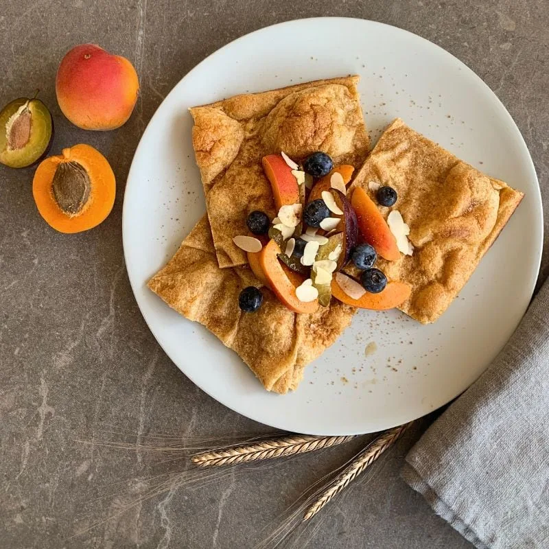 Ofenpfannkuchen aus Einkorn - Grundrezept. In quadrate geschnitten. mit Obst belegt. auf beigem Teller. daneben 2Aprikosen aufgeschnitten und Einkornähre.