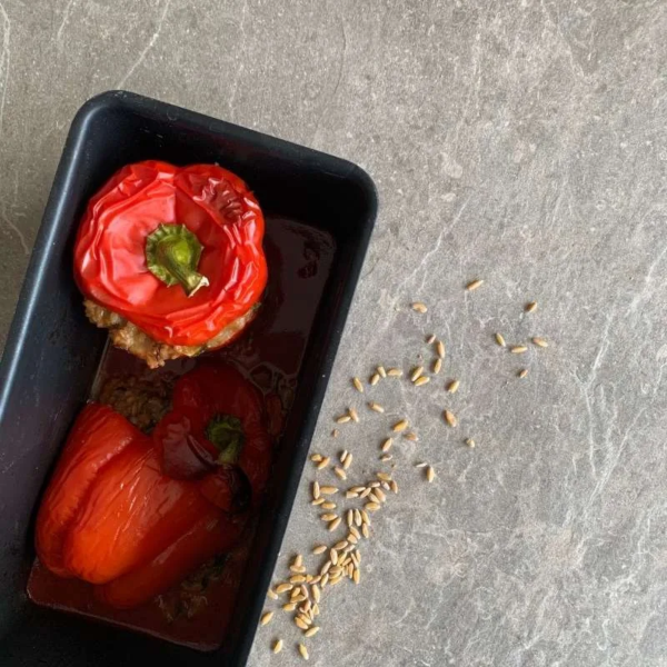 schwarze Backform auf einer Steinplatte. In er Form zwei rote gefüllte Paprika, frisch aus dem Ofen. Neben der Form liegen ein paar Getreidekörner