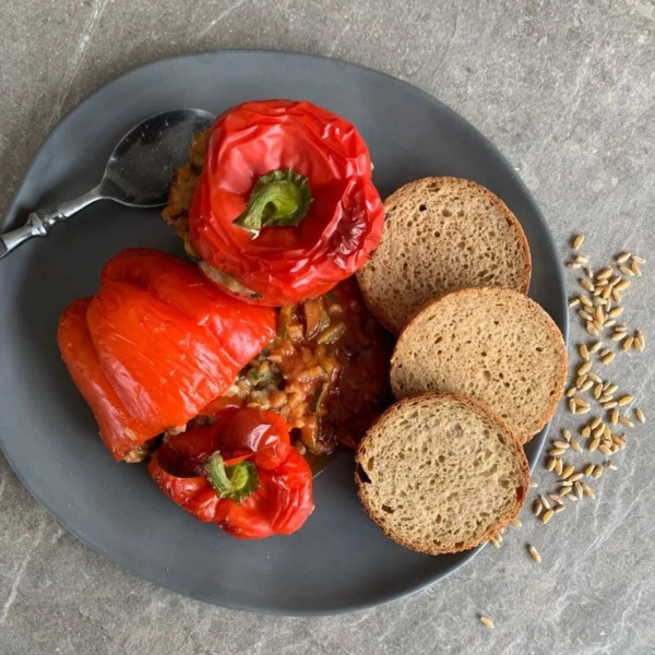 zwei rote gefüllte Paprika auf einem grauen Teller. Daneben liegen 3 runde Scheiben Urkornbrot und ein Löffel. neben dem Teller liege ein paar Getreidekörner
