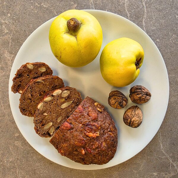 Auf dem Bild zu sehen ist eine taupe-farbene Steinplatte, auf welcher ein heller Teller zu sehen ist. Auf diesem Teller ist ein aufgeschnittenes Quittenbrot und auf der Schnittfläche des Brotes sind zudem Nüsse und Rosinen zu sehen. Rechts oben auf dem Teller, neben dem Quittenbrot liegen zwei reife Quitten und rechts darunter, ebenfalls neben dem Quittenbrot liegen drei geschlossene Walnüsse.