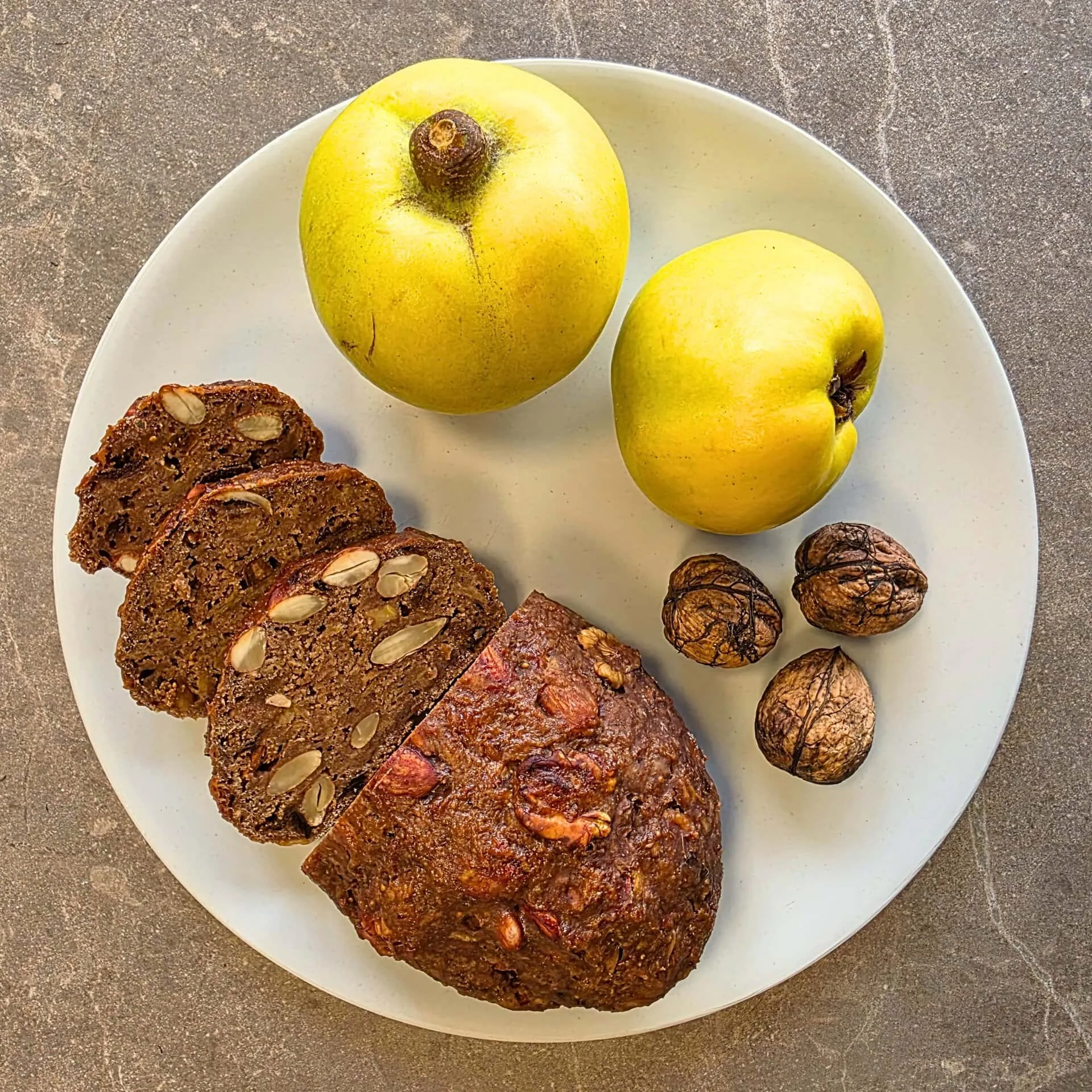 Auf dem Bild zu sehen ist eine taupe-farbene Steinplatte, auf welcher ein heller Teller zu sehen ist. Auf diesem Teller ist ein aufgeschnittenes Quittenbrot und auf der Schnittfläche des Brotes sind zudem Nüsse und Rosinen zu sehen. Rechts oben auf dem Teller, neben dem Quittenbrot liegen zwei reife Quitten und rechts darunter, ebenfalls neben dem Quittenbrot liegen drei geschlossene Walnüsse.