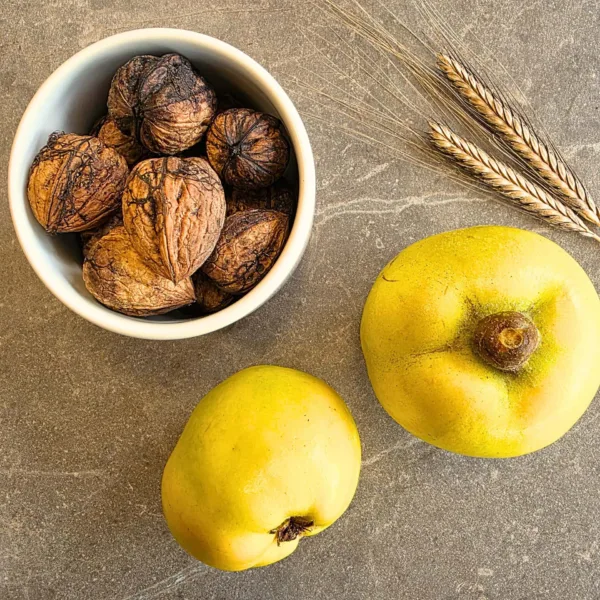 Zu sehen ist eine Steinplatte in der Farbe taupe. Darauf platziert sind 2 Quitten unten rechts, eine weiße kleine Schale von dem Unternehmen within mood gefüllt mit geschlossenen Walnüssen ist oben links zu sehen. Rechts oben sind zwei Ähren des Urgetreides Einkorn zu sehen.