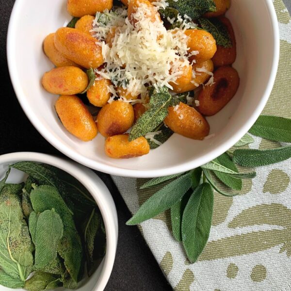 Auf dem Bild zu sehen sind zwei weiße Schälchen von dem Unternehmen within mood. welche auf einer dunkelgrauen arbeitsfläche stehen. Das grössere Schälchen ist oben zu sehen und halb angeschnitten. Darin befinden sich die orangenen Kürbis Gnocchi, welche mit in Butter geschwenktem Salbei und fein geriebenem Parmesan serviert wurden. Rechts unten ist ein beige frabenes Geschirrtuch zu sehen mit grünen Motiven, welche halb unter der großen Schale verschwindet. Rechts neben der großen schale auf dem Geschirrtuch befindet sich ein Zweig frischer Salbei, welcher zur Deko dient. In der kleinen Schale links unten sind die in Butter geschwenkten Salbeiblätter eingefüllt.