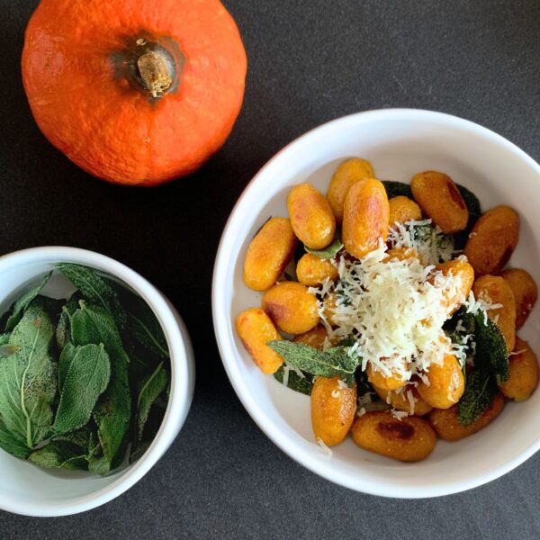 Auf der dunkelgrauen Arbeitsflächen sind drei Elemente zu sehen. Zum einen ein kleiner Hokkaido, welcher links oben halb ins Bild ragt. Mittig rechts ist eine etwas größere weiße Schale zu sehen, welche mit orangenen Kürbis Gnocchi gefüllt ist und in Butter geschwenktem Salbei und fein geriebenem Parmesan serviert wurde. Links daneben etwas unterhalb ist dasselbe Schälchen nur etwas kleiner zu sehen und wurde mit in Butter geschwenktem Salbei gefüllt.