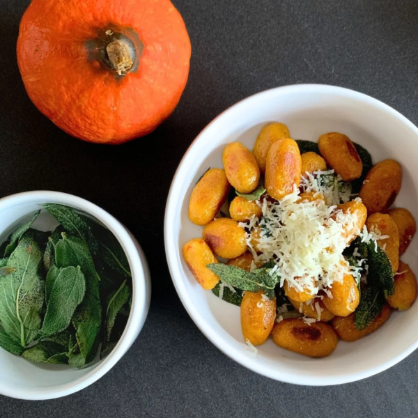 Auf der dunkelgrauen Arbeitsflächen sind drei Elemente zu sehen. Zum einen ein kleiner Hokkaido, welcher links oben halb ins Bild ragt. Mittig rechts ist eine etwas größere weiße Schale zu sehen, welche mit orangenen Kürbis Gnocchi gefüllt ist und in Butter geschwenktem Salbei und fein geriebenem Parmesan serviert wurde. Links daneben etwas unterhalb ist dasselbe Schälchen nur etwas kleiner zu sehen und wurde mit in Butter geschwenktem Salbei gefüllt.