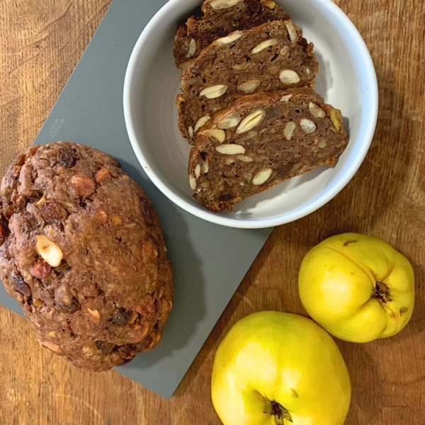 Der Hintergrund des Bildes ist eine alte Holzplatte, auf welcher ein graues Schneidebrett, sowie eine weiße Schale des Unternehmens within mood zu sehen sind. Das Schneidebrett liegt oben links schräg in der Ecke. Darauf platziert ist links das ganze Quittenbrot und rechts daneben die weiße Schale, welche mit Scheiben des Brotes gefüllt ist, Die scheiben liegen fächerartig in der Schale, mit der Schnittfläche nach oben und zu sehen sind Nüsse und Rosinen, welche im Brot enthalten sind. unten rechts neben dem Schneidebrett liegen zwei ganze Quitten.