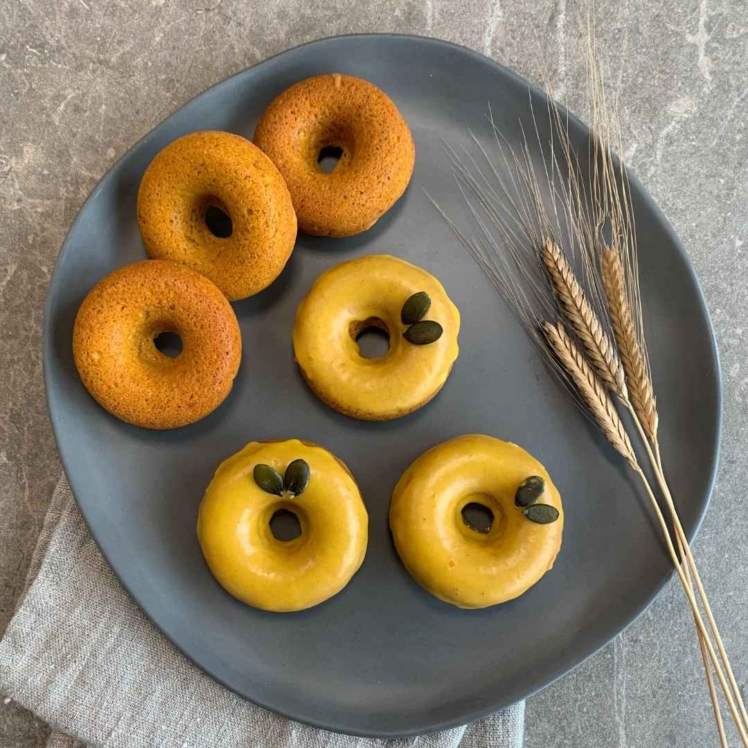 Sechs Kürbis-Einkorn-Donuts auf grauem Teller, teilweise glasiert, dekoriert mit Kürbiskernen und Getreideähren – herbstliche Backidee.