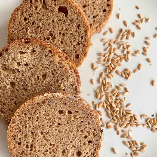 drei runde Scheiben Vollkornbrot BlueVelvet aus dem Glas liegen auf beigem Tisch. Daneben Getreidekörner