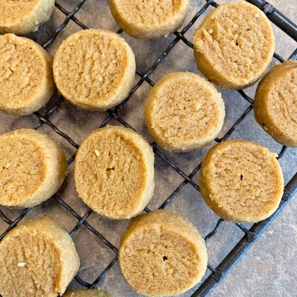 auf der Steinplatte in einem hellen taupe ton liegt ein Gitterrost zum auskühlen der runden und flachen Plätzchen. Die Plätzchen nennen sich Heidesand und sind leicht golden und rund. sie liegen auf dem Rost, welcher den Großteil des Bildes einnimmt. nur die untere rechte ecke ist frei.