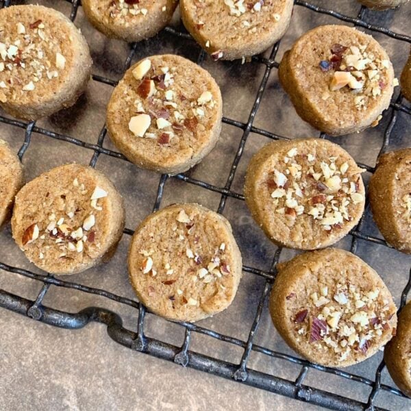 Auf der Steinplatte, welche einen hellen taupe ton hat liegt ein Gitterrost, auf welchem kleine runde Plätzchen auskühlen. diese sind mit klein gehackten Nüssen verziert. Die Plätzchen sind flach und sind leicht braun bzw golden. die Plätzchen und das Gitter nehmen den Großteil des Bildes ein. nur die linke untere Fläche ist frei.