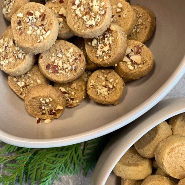 Auf dem Bild sind zwei creme farbene Schüsseln und ein Thujazweig zu sehen. Die größere Schüssel ist angeschnitten zu sehen und nimmt den großteil des bildes ein. Sie kommt aus der linken oberen ecke. In der schüssel liegen Plätzchen mit klein gechackten Nüssen als Topping. Darunte in der rechts unteren ecke ist eine kleinere Schüssel angeschnitten, in welcher dieselben kleinen runden Heidesand-Plätzchen liegen, allerdings ohne Nüssen on top, also ganz klassisch. in der linken unteren Ecke ist der Thujazweig zu sehen.