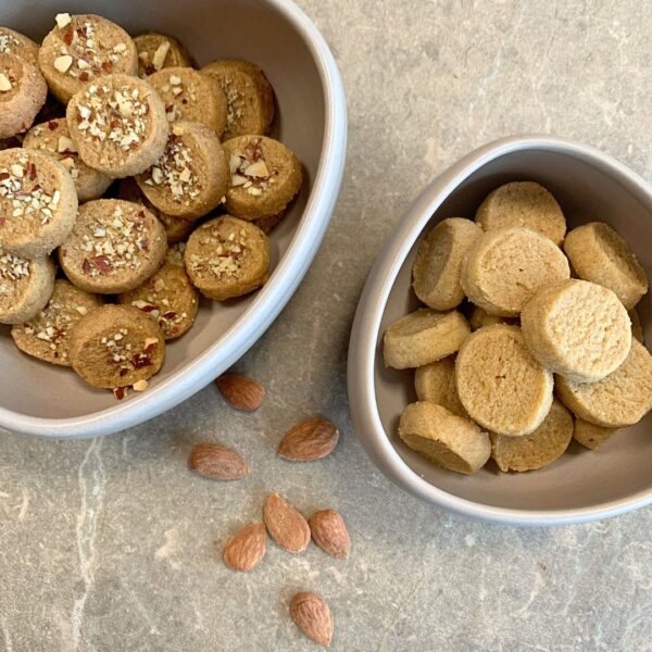 auf der steinplatte mit einem hellen taupe ton stehen zwei Schüsseln in einem creme ton und ein paar Mandeln. Die größere Schüssel ist angeschnitten in der linken oberen ecke zu sehen. darin befinden sich runde flache Plätzchen, welche mit klein gehackten nüssen verziert sind. die kleiner schüssel ist rechts angeschnitten in der mitte des bildes zu sehen. in ihr befinden sich dieselben plätzchen allerdings ohne die Nussverzierung. die plätzchen sind golden und sehen sehr buttrig aus. unterhalb zwischen den Schüsseln liegen sieben verstreute Mandeln zur Deko aus.