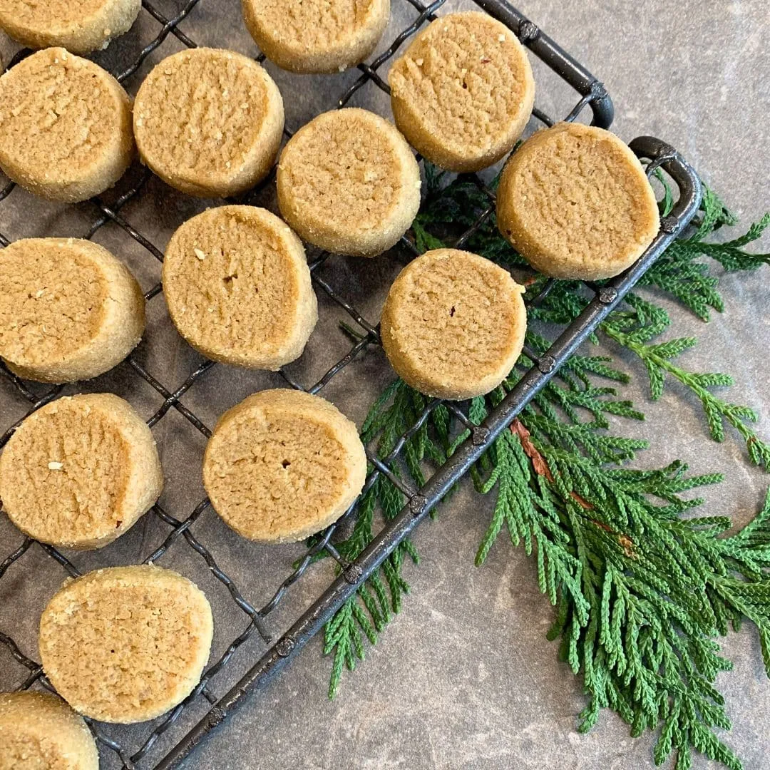 runde Weihnachtsplätzchen, die Heidesand heißen, liegen auf einem Gitterrost verteilt zum Auskühlen. Die Plätzchen sind golden und rund. Das Gitter mit den Plätzchen ist auf der linken seite des bildes zu sehen. rechts ragt ein Thujazweig unter dem gitter hervor. Alle Elemente liegen auf einer hellen creme farbenen Steinplattte