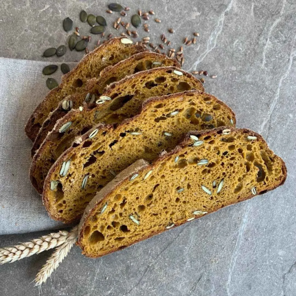 Fünf Scheiben Brot vom Herbstwald Brot mit Kürbis liegen aufgefächert übereinander auf einer grauen Platte und einem Stück Tuch. Danben 2 Getreideähren und ein paar Kürbiskerne
