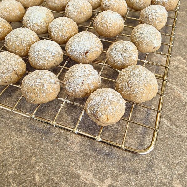 Zu sehen ist auf einer hellen Steinplatte ein goldener Gitterrost, welcher den Großteil des Bildes einnimmt. Der Rost wird von der linken oberen Ecke des Bildrandes angeschnitten und ist somit nur teilweise zu sehen. Auf dem Rost befinden sich helle rund Plätzchen, welche mit Puderzucker bestreut sind.