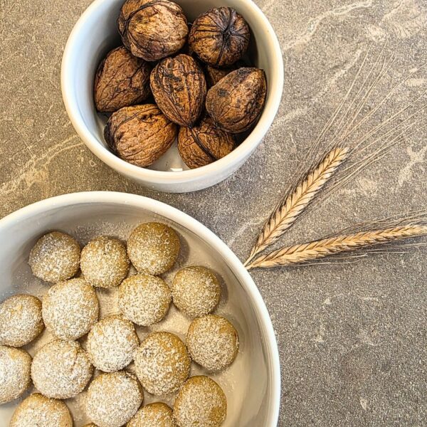 Zu sehen sind zwei weiße Schalen auf einer Steinplatte. Die Steinplatte hat die Farbe beige und ist sehr natürlich. Das Licht ist hell und allgemein ist das Bild in warmen hellen und natürlichen Farben gestaltet. Die weißen Schalen haben zwei verschiedene Größen. Die kleiner Schale ist oben zu sehen un leicht angeschnitten. Darin befinden sich sieben ungeöffnete Walnüsse. Darunter in der linken Ecke ist angeschnitten die größere weiße Schale zu sehen. In ihr befinden sich helle kleine und runde Plätzchen, welche mit Puderzucker bestreut sind. rechts sind zwei Ähren des Einkorns zu sehen.