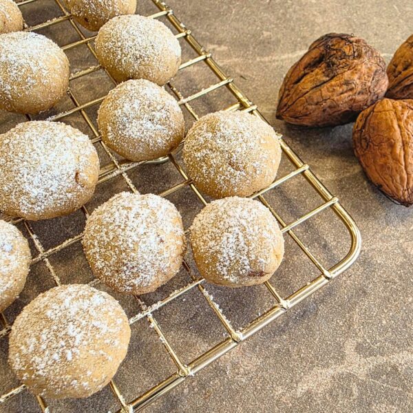 Zu sehen ist ein goldenes Gitterrost welches die linke seite des Bildes einnimmt und mit der ecke in die rechte ecke des Bildes geht. Darauf befinde sich helle runde Plätzchen, welche mit Puderzucker bestreut sind. Daneben, ebenfalls auf der hellen Steinplatte, befinden sich drei ungeöffnete Walnüsse, welche teilweise vom rechten Bildrand angeschnitten sind. Das Bild ist von der Farbgebung hell, freundlich und warm.