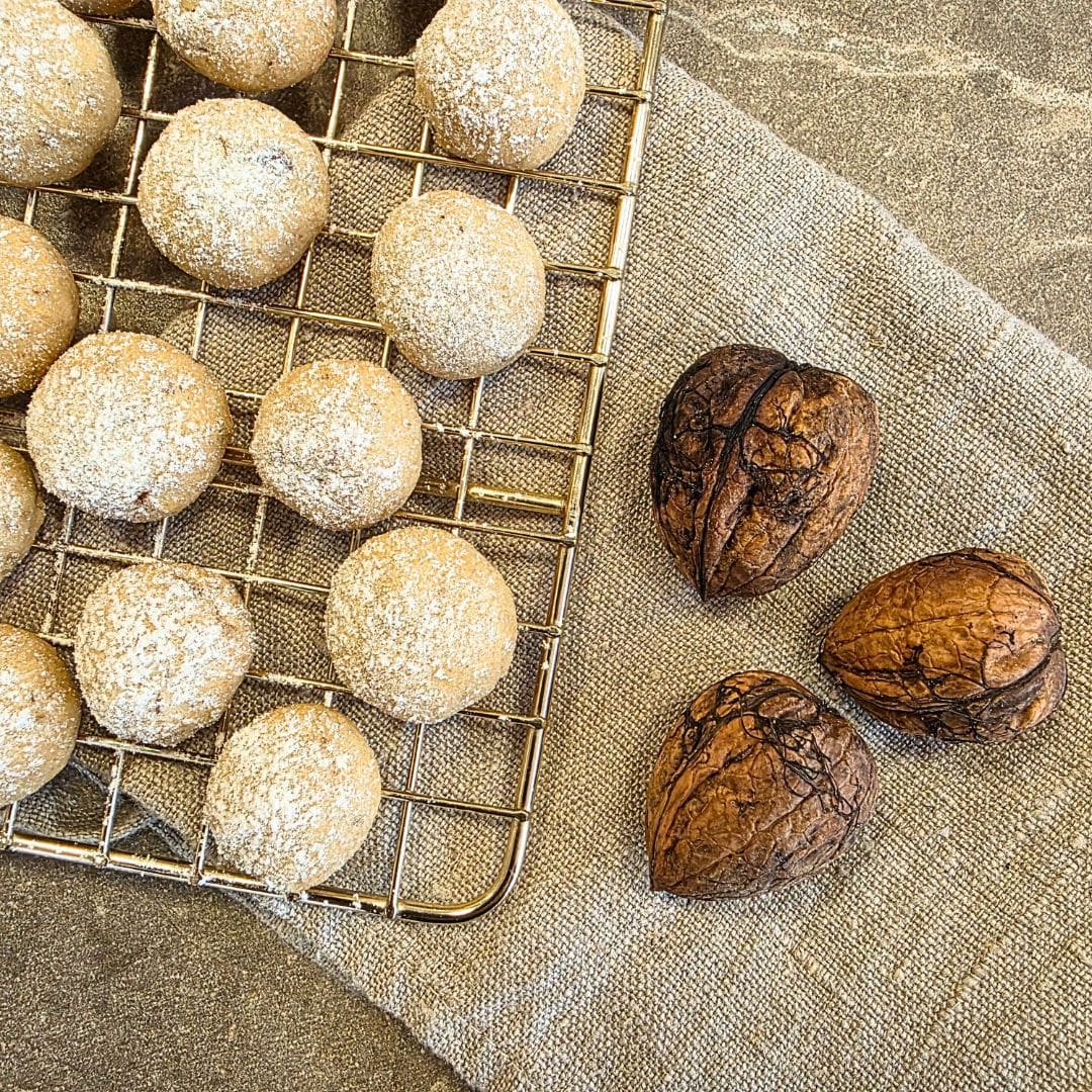 Zu sehen ist ein goldener Rost zum Auskühlen von Backwaren. Der Gitterrost liegt auf einer Steinplatte, welche beige ist. Außerdem liegt unter dem Rost ein beiges Leinengeschirrtuch, welches zusammengefaltet ist und halb unter dem Rost liegt und zur anderen hälfte nicht. Der Rost liegt in der linken oberen Ecke und nimmt fast die Hälfte des Bildes ein. Das Geschirrtuch liegt schräg im Bild und nimmt den Großteil der Arbeitsfläche ein. Auf dem Teil des Geschirrtuches, welches nicht unter dem Rost liegt, liegen drei ungeöffnete Walnüsse. Auf dem goldenen Gitterrost liegen Plätzchen, welche hell, rund und klein geformt und gebacken sind. Sie sind zudem mit Puderzucker bestreut.