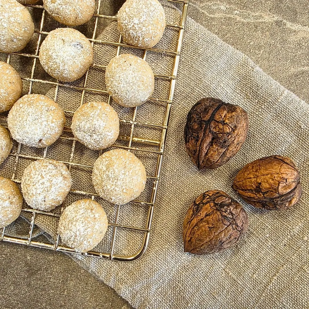 Zu sehen ist ein goldener Rost zum Auskühlen von Backwaren. Der Gitterrost liegt auf einer Steinplatte, welche beige ist. Außerdem liegt unter dem Rost ein beiges Leinengeschirrtuch, welches zusammengefaltet ist und halb unter dem Rost liegt und zur anderen hälfte nicht. Der Rost liegt in der linken oberen Ecke und nimmt fast die Hälfte des Bildes ein. Das Geschirrtuch liegt schräg im Bild und nimmt den Großteil der Arbeitsfläche ein. Auf dem Teil des Geschirrtuches, welches nicht unter dem Rost liegt, liegen drei ungeöffnete Walnüsse. Auf dem goldenen Gitterrost liegen Plätzchen, welche hell, rund und klein geformt und gebacken sind. Sie sind zudem mit Puderzucker bestreut.